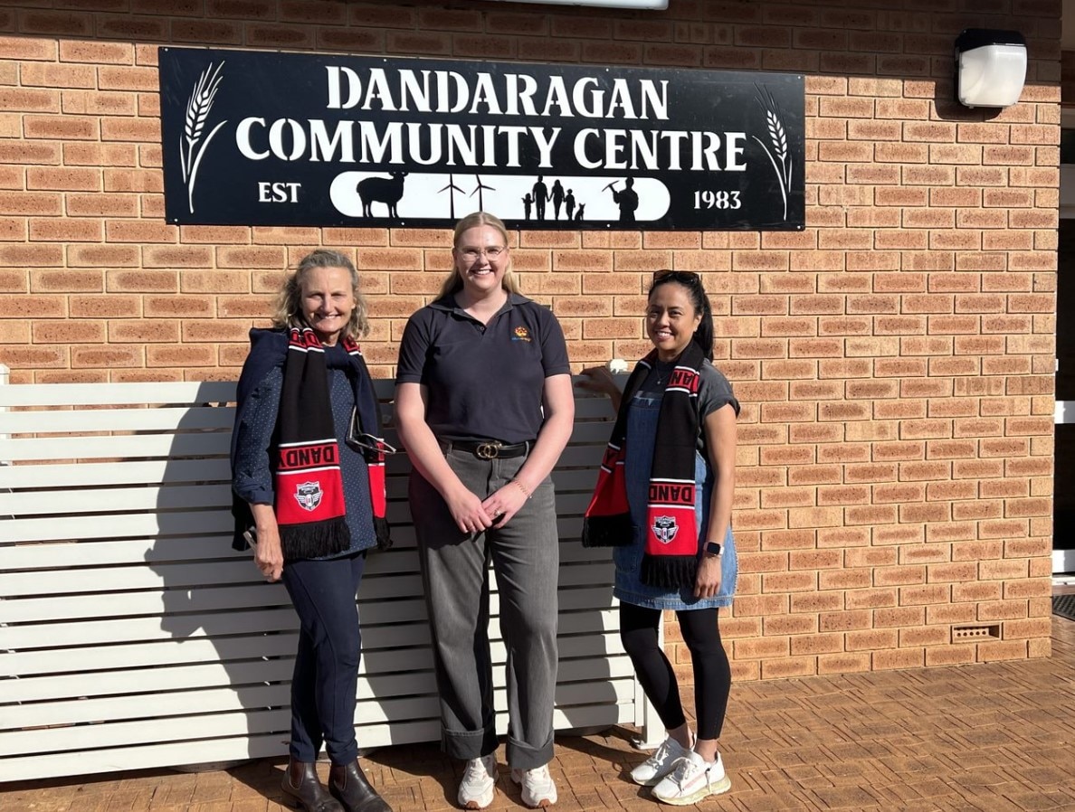 The image shows three people standing outside the Dandaragan Community Centre. A sign displaying the centre's name is mounted on the wall behind them.